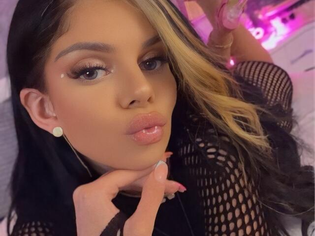 IvyRaven with glossy pink lips, blonde and black hair, fishnet top, holding her chin, wearing round earrings, purple neon lights in the background, and a pink ring.