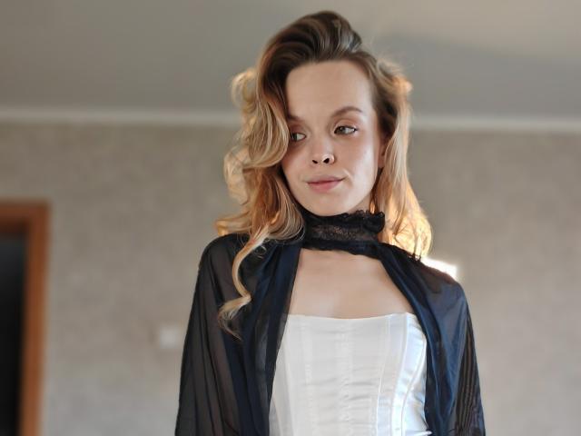 Sinteas, met golvend blond haar, draagt een zwarte, doorschijnende robe over een witte corset, heeft een subtiele glimlach, zachte bruine ogen en een vriendelijke uitdrukking..