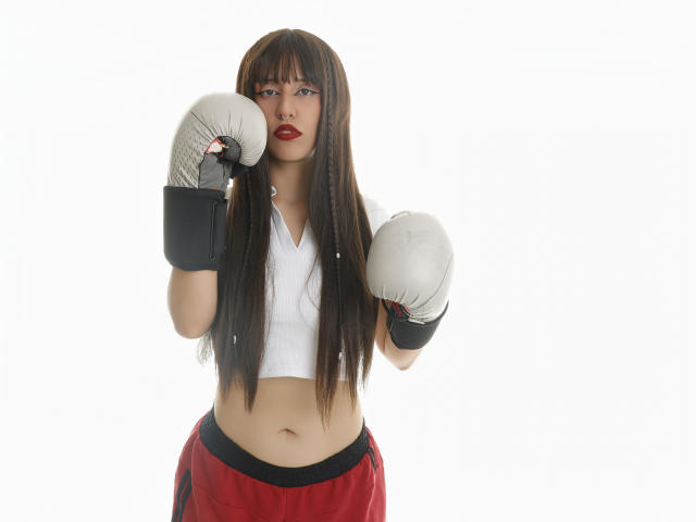 Kaylin Krof, com cabelos castanhos longos, top branco cropped, calças de boxe vermelhas e luvas de boxe brancas, em pé com os punhos levantados, com uma expressão séria e batom vermelho..