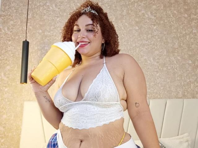 YeraCarey with curly brown hair, wearing a white lace halter top, sipping a yellow drink with a white straw, showing her big tits and yellow thong.