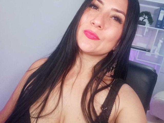 KatyaScott, com cabelos longos e pretos, batom rosa e blusa de renda preta, fazendo beicinho, mostrando os seios, usando brincos pendurados, em um selfie..
