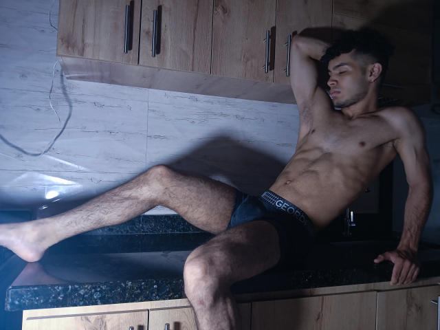 ZafkielBoby, muscular and hairy, sits on a kitchen counter in dark underwear, one leg up, arm behind head, eyes closed, with a relaxed, sexy, and confident expression.