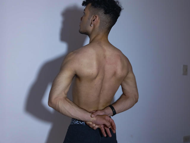 ZafkielBoby's back is to the camera, showing a muscular, shirtless body with dark curly hair, ear piercing, and hands on his ass. Shadow on white wall.