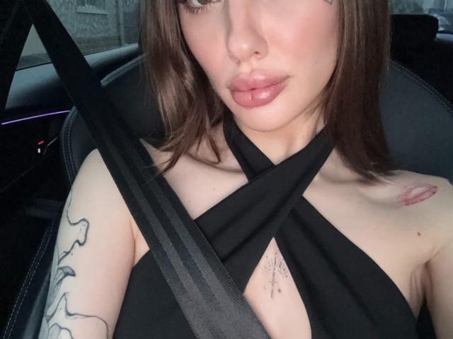 SensualMila with brown hair, wearing a black halter top, seatbelt across chest, showing tattoos on left arm and right chest, pouty pink lips, car interior, subtle makeup.