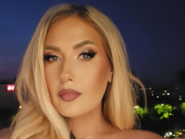 OndinaNoir has blonde hair, green eyes, and full pink lips with dark eyeliner. Her ass is not visible, just her face and shoulders.