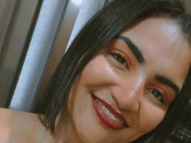 SusanaDiaz has shiny brown skin, thick black eyebrows, and a big smile with red lipstick showing her teeth. She's got black hair and a cute face.