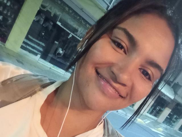 SusanaDiaz has a cute smile, brown eyes, and earbuds in her dark brown hair. She's wearing a white shirt with a brown jacket, showing her shiny pink lips.