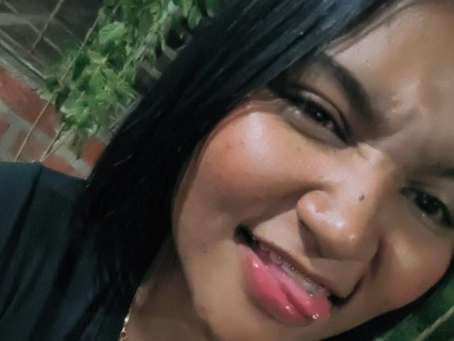SusanaDiaz's face, black hair, shiny wet skin, slightly open mouth with tongue out, small moles, pink lips, dark eyes, wearing black top, green vine in background.