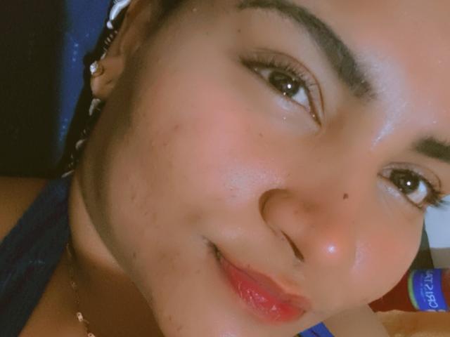SusanaDiaz's face, showing a slight smile with red lips, glossy skin, dark eyebrows, brown eyes, and a small beauty mark on the right cheek. She wears a blue top.