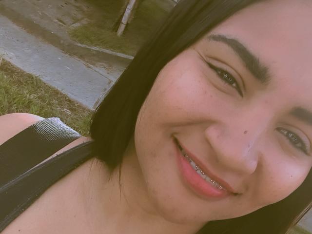 SusanaDiaz with black hair, thick eyebrows, and a cute smile, showing metal braces on her teeth, wearing a black tank top. Her skin is light, and she's outdoors.