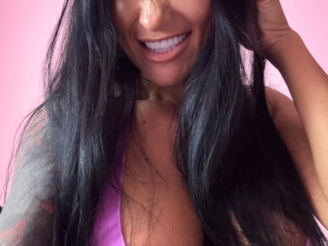 AndraD has long black hair, smiling with white teeth, wearing a purple top, showing cleavage, and touching her hair with her right hand.