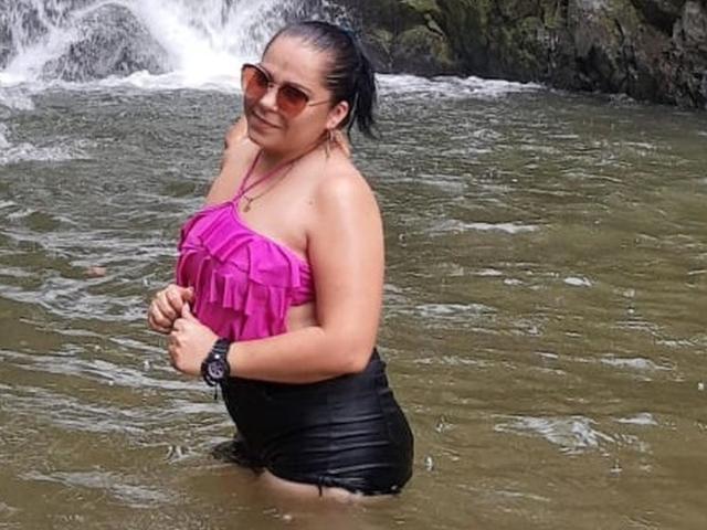 DulceHunny has wet black shorts, a pink ruffled top, and brown sunglasses. She's wading in water, hands near her chest, with a black wristwatch on.