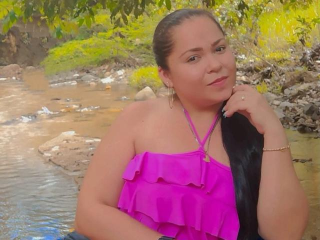 DulceHunny with long black hair, pink ruffled top, hoop earrings, and gold bracelet, smiling near a rocky stream, right hand on chin. Her tits are slightly visible.