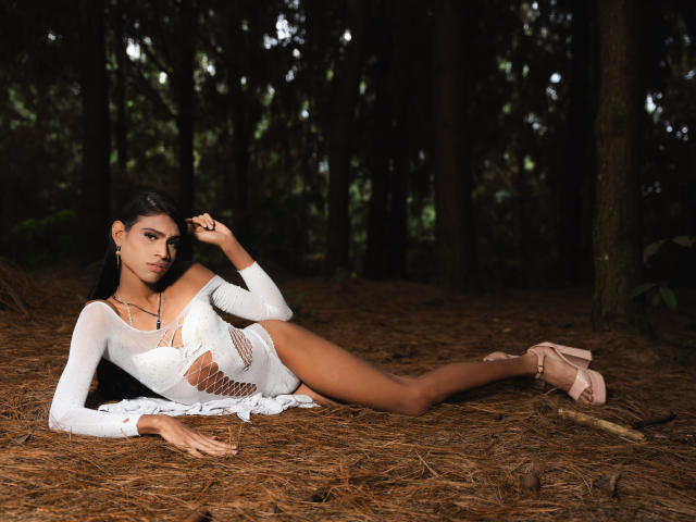 LissyAndrade laying in a forest, wearing a white, cut-out dress, showing her tits, with her leg raised, in pink heels, dark hair, and a seductive look, on pine needles.