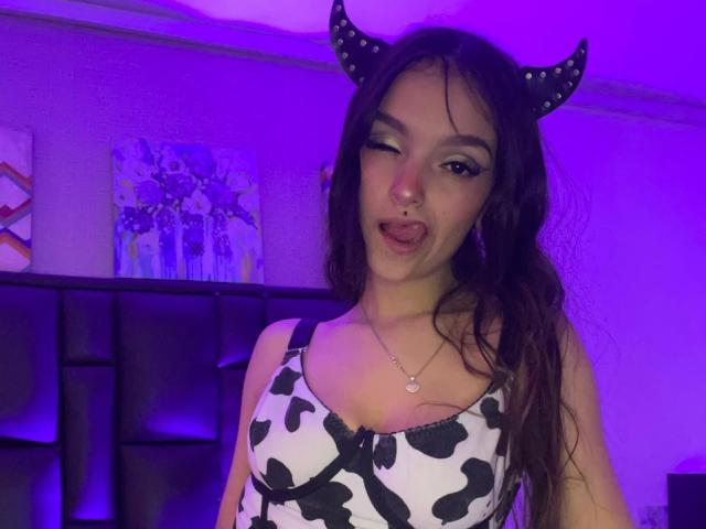 MarietteJones with long brown hair, black and white cow print dress, devil horns, winking, tongue out, nose ring, purple lighting, necklace, looking at camera, in a bar setting.