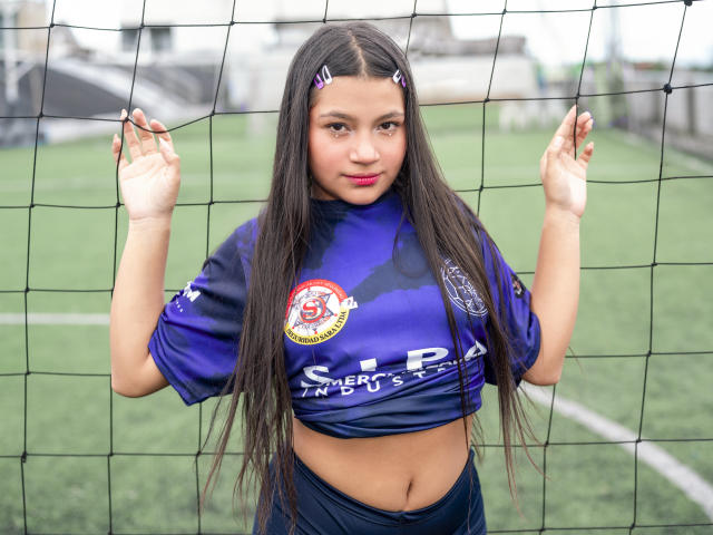 Daniela Lohan, die vor einem Fußballtor steht, hat langes, schwarzes Haar, ein violettes Fußballtrikot und dunkle Hosen. Sie zeigt ihren Bauch und hält das Netz mit beiden Händen..