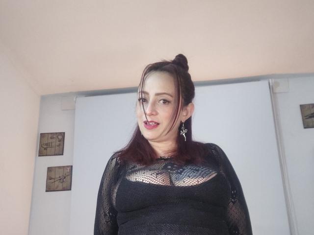 CatalinaMonyque with pale skin, red hair in a bun, black mesh top showing her tits, and red lipstick, standing in front of a white wall, looking at the camera.
