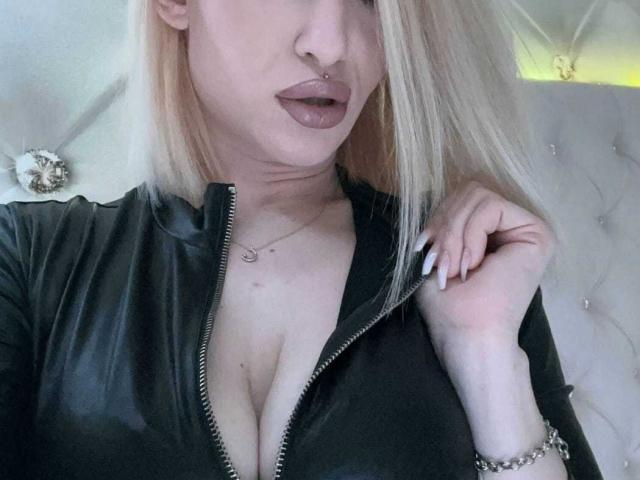 LanaSladkih, con i capelli biondi, indossa una giacca di pelle nera con zip che lascia intravedere il décolleté, ha un piercing al naso e rossetto viola, tiene la zip, ha le unghie bianche, un braccialetto d'argento e un tatuaggio discreto sul petto..