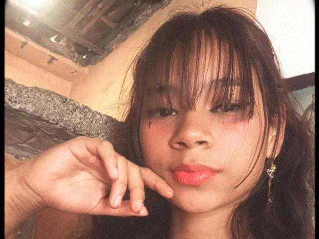 GabrielaJam with messy black hair, red lipstick, small tattoo under right eye, hand near mouth, brown eyes, earrings, soft smile, slightly dirty skin, casual selfie.