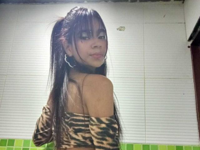 GabrielaJam with long black hair in a ponytail, wearing a tiger-print off-shoulder top, looking over her shoulder with a slight smile, showing her bare back and small ass.