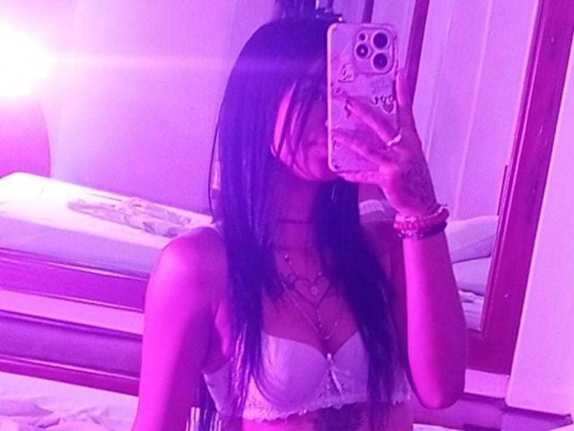 GabrielaJam with long brown hair, wearing a white lace bra, taking a mirror selfie in purple-lit room, covering face with phone, visible tits, bracelets on right wrist.