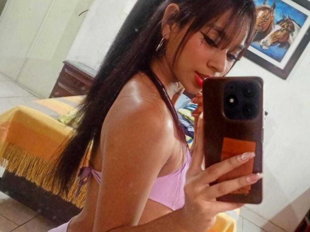 GabrielaJam with long black hair, pink bra, and red lipstick, holding a phone, taking a mirror selfie, showing her small tits and slim body, in a bedroom.