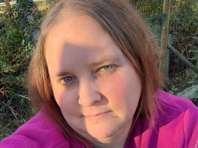 Kiramaus88-hot has a chubby face, light green eyes, reddish-brown shoulder-length hair, and a pink nose piercing. Wearing a pink shirt, she's outside in the sunlight.