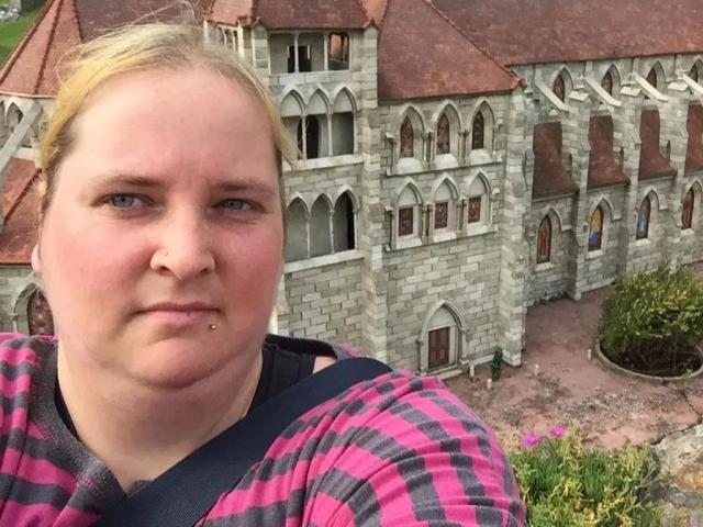Kiramaus88-hot has pale skin, blonde hair, and a pimpled lower lip. She's wearing a pink and black checkered shirt, with a black strap over her shoulder.