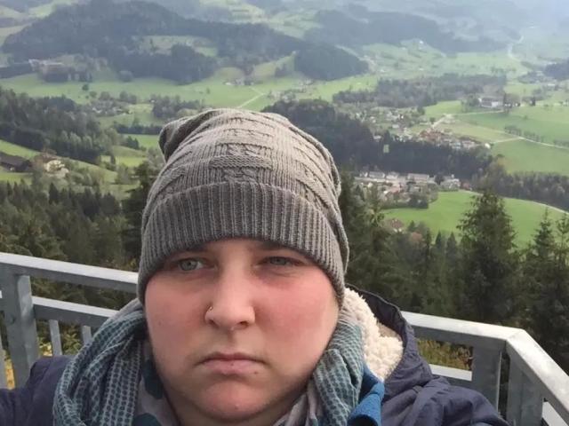 Kiramaus88-hot with a chubby face, blue eyes, nose ring, gray knit beanie, blue jacket, and gray scarf, looking serious. Background is a green, hilly landscape.