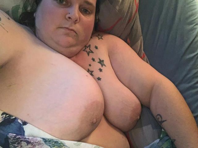 Kiramaus88-hot has large, saggy tits with star tattoos across her chest, a nose ring, and a tattoo on her right arm, lying on a bed, looking at the camera.