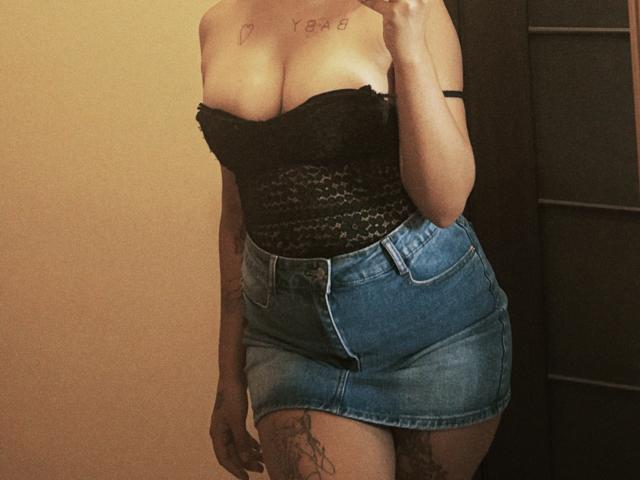 Kametia with big boobs in a black lace strapless top and a blue denim skirt, showing tattoos on her chest and thigh, taking a mirror selfie.