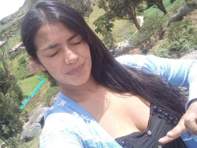 ArianithaXox with long black hair, eyes closed, wearing a blue cardigan and black top showing her tits, pointing at her chest in a sunny, green outdoor setting.