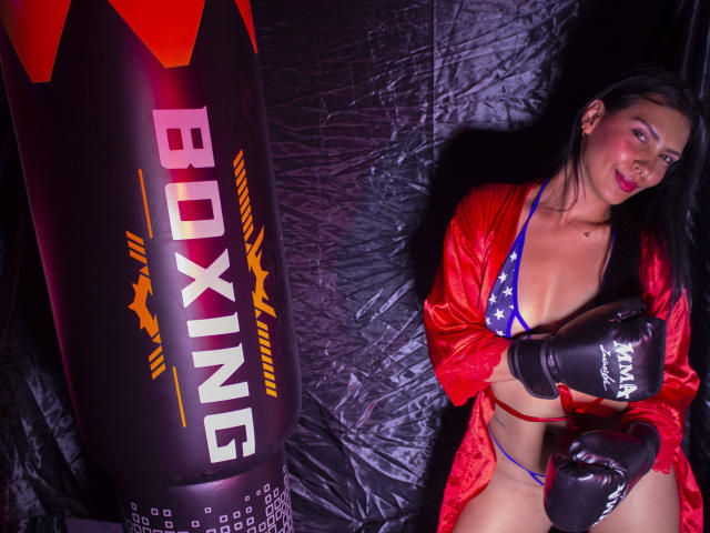 Alyson Stone, con i capelli neri, gli occhi rossi e un sorriso seducente, indossa un bikini con stelle, un vestito rosso e dei guantoni da boxe neri, ed è accanto a un sacco da boxe nero..