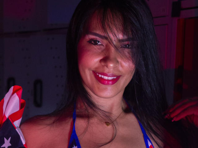 AlysonStone with dark hair, red lipstick, and a smile, holding a flag on her shoulder, wearing a blue halter top, and a gold necklace.