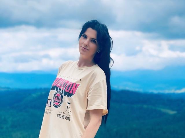 VioletaStyle with long black hair, smiling, wearing a beige T-shirt, hands in pockets, blue sky and green hills in the background, standing, looking slightly to the left.