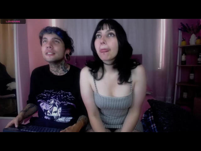 TwoNightmares with blue-tinted hair and tattoos, wearing a black shirt with a wolf print, sits next to TwoNightmares with black hair and a checkered dress, looking up.