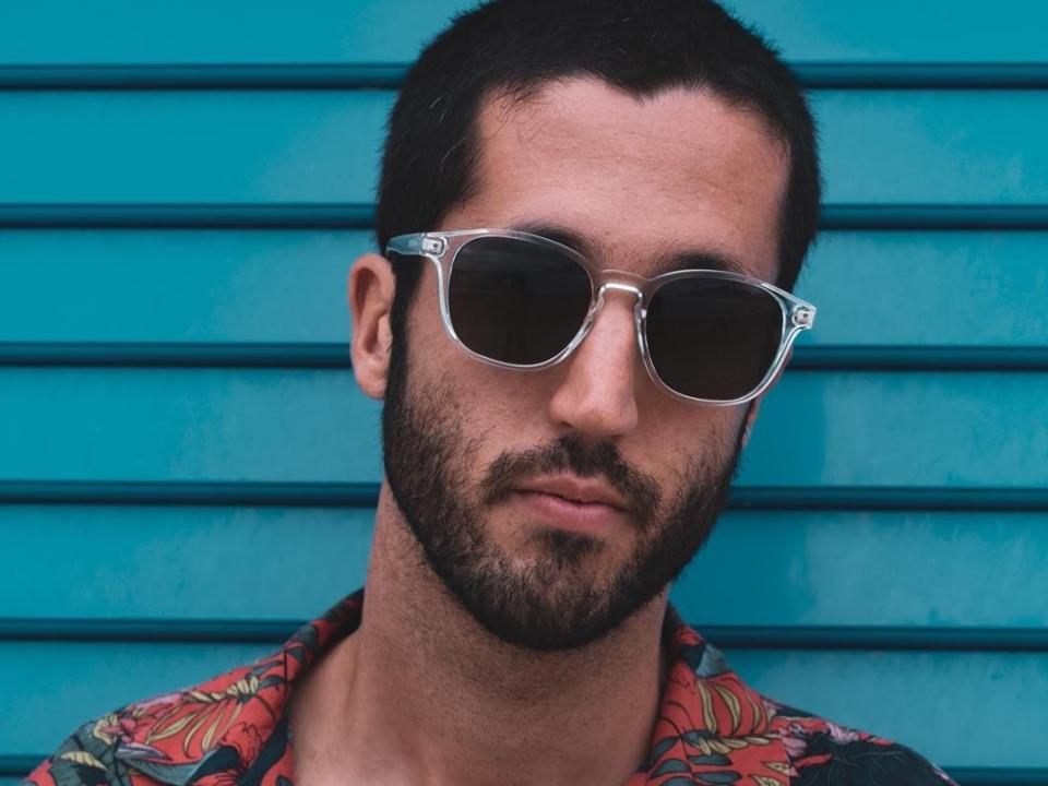 Lucas69s has a beard, short dark hair, and is wearing clear plastic aviator sunglasses with a red and blue floral shirt. His face is serious and centered.