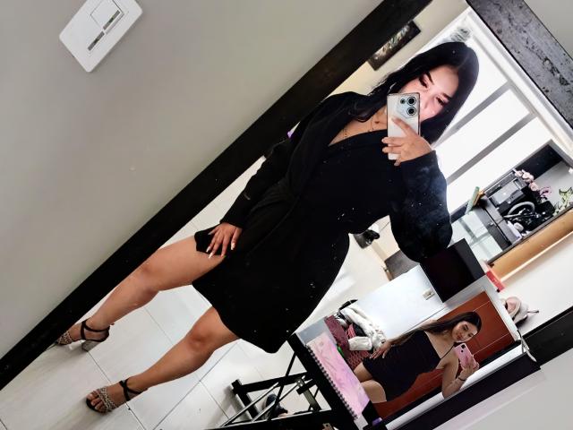 CamilaBrownXx with black dress, showing thigh, holding phone, wearing strappy heels, mirror selfie, reflection, long black hair, fair skin, medium breasts, black nails, white wall, black trim.