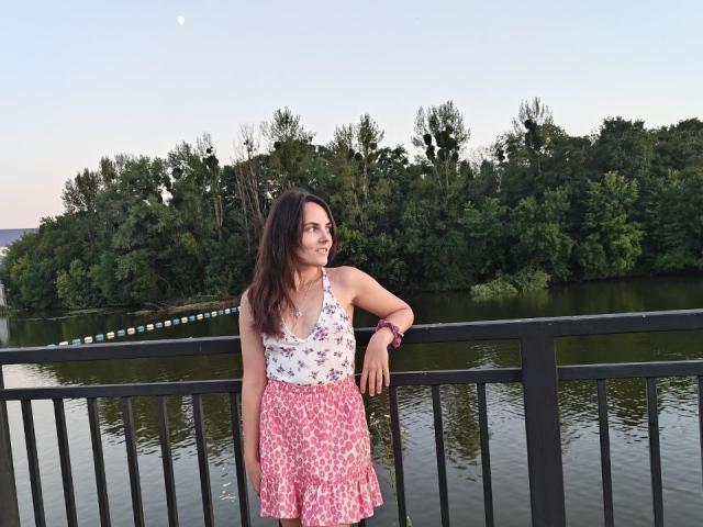 AlinaBoss met lang bruin haar, een witte top met bloemenprint, een roze rok met bloemenprint, leunt tegen een zwarte reling, glimlacht naar de maan in de avondhemel, draagt een roze armband en staat bij kalm water..