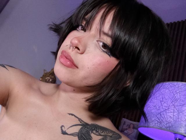 NatashaHottson with black bob, nose ring, and winged eyeliner, topless, showing a tattoo on her right shoulder, purple lamp in background, slightly pouty lips.