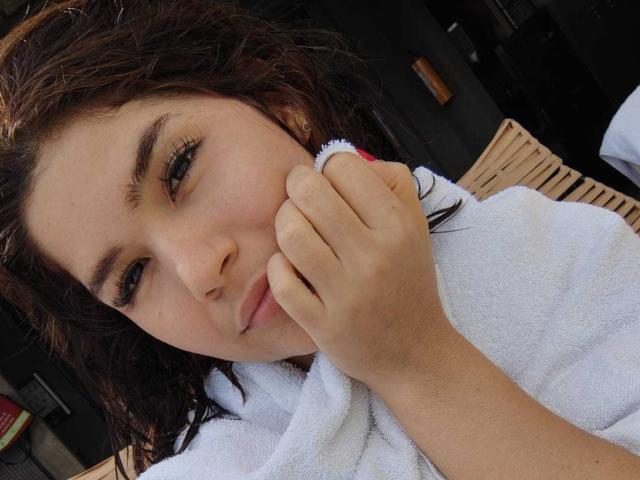 ScarlletZhen with wet brown hair, holding a white towel to her face, looking down with slightly parted lips, wearing subtle makeup, with a small pink ring on her right hand.