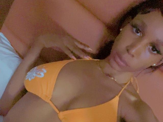 SoftSuga with medium brown skin, wearing an orange bikini top with a white flower, has small tits and a gold necklace, lying down with a hand on her chest.