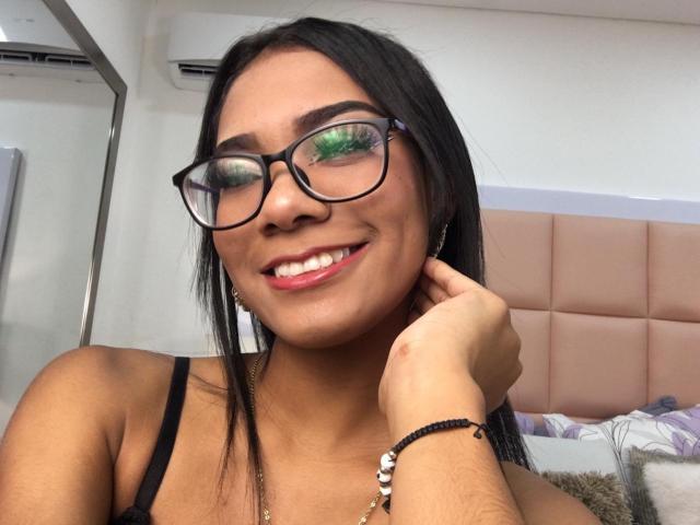 SusanFarrer with dark hair, glasses, and pink lipstick, smiling, showing her teeth, wearing black straps, hand on neck, green reflection in glasses, bracelet on wrist.