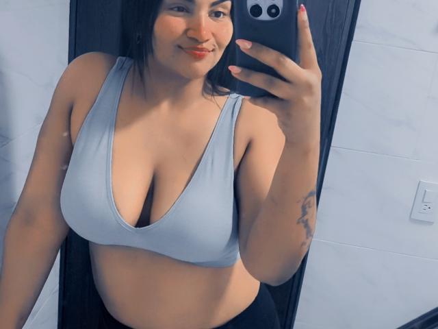 SusanaDiaz with medium tits in a light gray sports bra, holding a phone, smiling, black hair, tattoo on right arm, black pants, taking a mirror selfie.