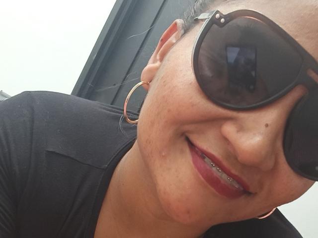SusanaDiaz with dark sunglasses, red lipstick, gold hoop earrings, black shirt, smiling with visible teeth, freckles on her face, slightly tilted head, close-up selfie.