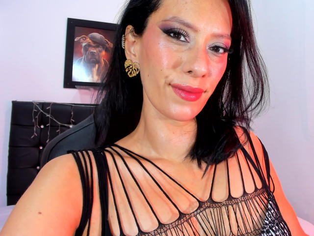 PriscilaSamis with black hair, wearing a strappy black dress, showing her cleavage, pink lipstick, and gold earrings, smiling at the camera. Boobs visible through dress.