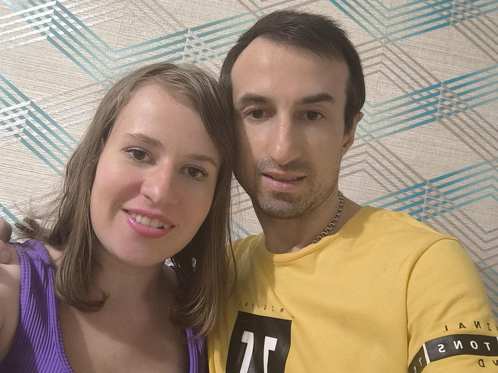 LallyJodie with brown hair, pink lipstick, and purple top, and LallyJodie with short dark hair, beard, yellow shirt, and necklace, smiling at the camera.