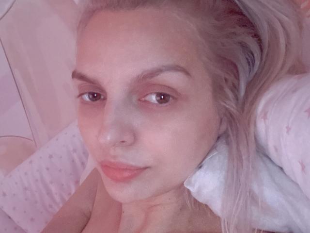 DouceAmelia's face with light blonde hair, brown eyes, and pinkish skin, lying on a pillow, looking up with a slight smile, in a soft, pink-lit image.