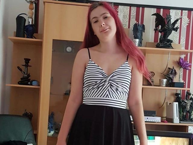Spielzeuglena-hot with red hair, wearing a striped black and white top, and a black skirt, standing in a room with wooden shelves and various small statues.