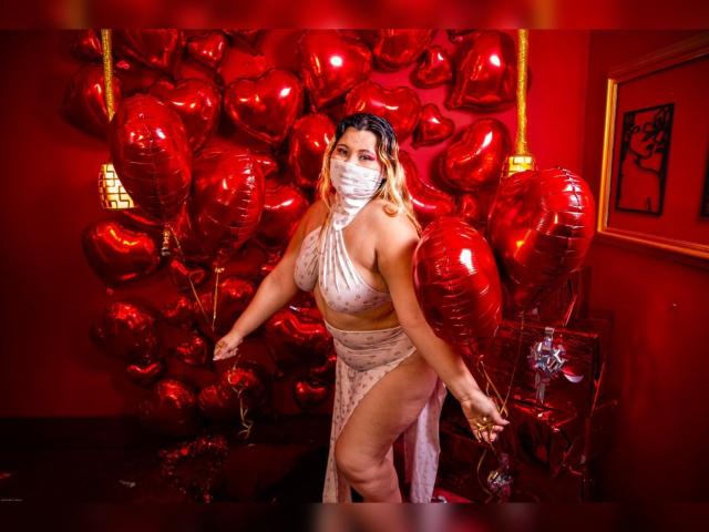 SalmaAzaman in a shiny, white, halter dress with a high slit, holding red heart balloons, wearing a white mask, blonde hair, red walls, and a gold frame in the background.